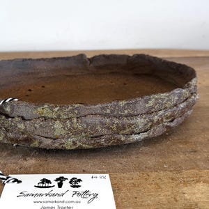 May include: A stack of three oval-shaped, dark brown ceramic bonsai pots with a rough, textured exterior and a smooth, dark interior. A white tag attached with black and white twine displays the text Samarkand Pottery, www.samarkand.com.au, and James Tranter.
