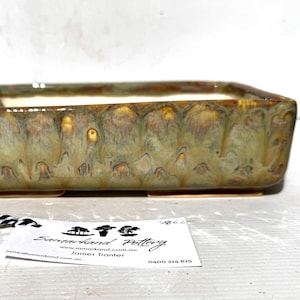 Puede incluir: Jardinera rectangular de cerámica con acabado vidriado en tonos verdes, marrones y amarillos. La jardinera tiene una superficie ligeramente texturizada y un interior claro. Se adjunta una etiqueta con el texto "Samarkand Pottery".