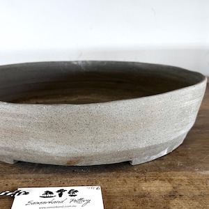 May include: A shallow, oval-shaped ceramic bonsai pot with a textured, speckled light grey finish. The exterior surface shows subtle variations in tone with faint reddish-brown markings. A white tag with the text Samarkand Pottery is attached.