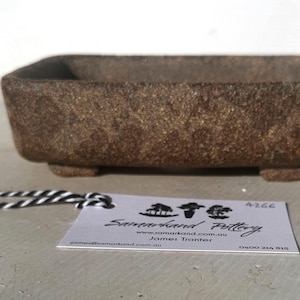 May include: A rectangular, brown stone planter with a textured surface, resting on four small feet. A tag with the text "Samarkand Pottery" and contact information is attached.