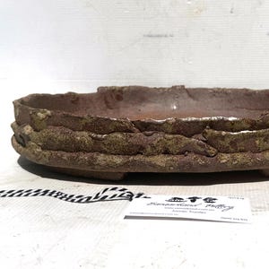 May include: A handcrafted, oval bonsai pot. The pot is a deep reddish-brown colour with textured, layered sides and a rough, uneven rim. The pot has a weathered appearance, suggesting it is made of ceramic or clay. A small tag is visible.