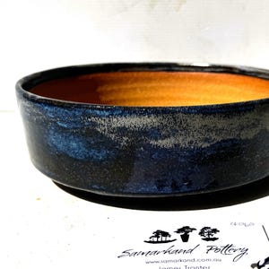 May include: A handcrafted ceramic bowl with a wide, shallow form. The exterior is a dark blue with a textured, mottled finish, and the interior features a warm orange-yellow glaze. This bowl is suitable for serving food or as a decorative item.
