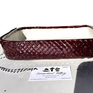 May include: A rectangular ceramic dish with a textured, deep burgundy exterior and a white interior. The dish has a tag attached with the text "Samarkand Pottery" and contact information. The dish is approximately 25 cm long.
