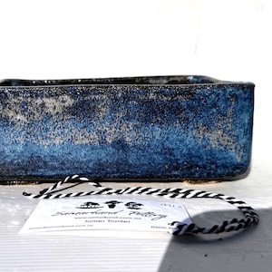 Könnte beinhalten: Rechteckiger Bonsai-Topf mit strukturierter, blau-grauer Glasur. Der Keramiktopf hat einen dunklen Rand und steht auf kleinen Füßen. Vor dem Topf befindet sich ein Etikett mit schwarz-weißem Faden.