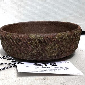 May include: A handcrafted, round, brown pottery bowl with a textured, bark-like exterior. The bowl has a rough, earthy appearance, with hints of green and red hues. A tag with black and white string is attached.
