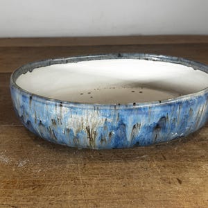 May include: A shallow, oval ceramic bowl featuring a smooth, solid white interior and a textured exterior with a reactive glaze in shades of deep blue, light blue, and grey, creating a vertical drip pattern around the outer rim.