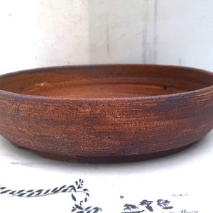 May include: A handmade, round, brown ceramic bowl with a rustic, textured finish. The bowl has a wide, shallow design, suitable for serving or display. The exterior has a dark brown colour, while the interior is a lighter shade of brown. The bowl is approximately 5 cm tall.