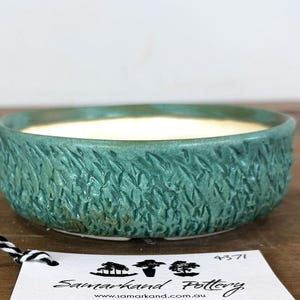 May include: A round, shallow ceramic bowl with a textured, teal-green exterior featuring a repeating leaf-like pattern. The interior of the bowl is smooth and off-white. A white tag with black text reading Samarkand Pottery is attached by a string.