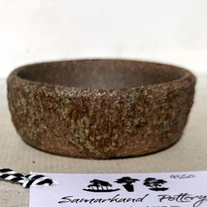 May include: A round, low-profile ceramic bowl with a rough, textured exterior in shades of brown and muted green. A white tag is attached with black and white twine, featuring the text Samarkand Pottery and three small tree illustrations.