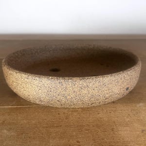 May include: A shallow, round ceramic bowl with a speckled, textured tan and brown finish. The interior base features a small, circular drainage hole, indicating its function as a planter or bonsai pot with a thick, sturdy rim and walls.