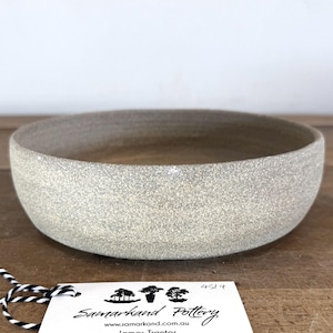 May include: A round, shallow ceramic bowl with a speckled, light beige matte finish. Attached to the bowl is a white rectangular tag featuring the text Samarkand Pottery, www.samarkand.com.au, James Tranter, and the number 4514 in black ink.