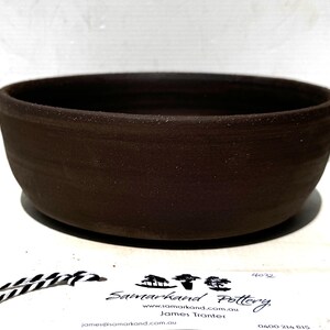 May include: A dark brown, circular ceramic bowl with a matte finish and subtle horizontal ridges along the exterior wall. A white tag is positioned in front, featuring the text Samarkand Pottery, a website URL, and contact information.