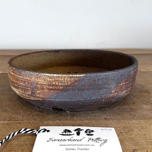 May include: A shallow, round ceramic bonsai pot with a textured, dark brown and burnt orange crackle glaze finish. A white rectangular tag is attached with black and white string, featuring the text Samarkand Pottery, James Tranter, and 4510.