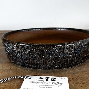 May include: A round, shallow ceramic bowl with a dark, heavily textured exterior featuring a rough, craggy finish in shades of brown and black. The interior is smooth and matte brown. A white tag with black text reads Samarkand Pottery.