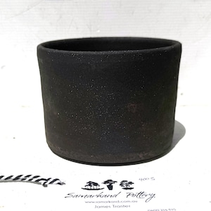 May include: A handmade, cylindrical black pottery vessel with a textured surface. The open-topped container is a dark charcoal colour and sits on a white surface. The base is slightly darker than the body. The pottery is handmade.