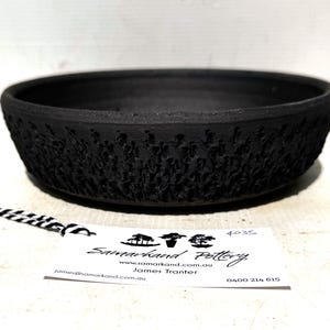 May include: A shallow, circular black ceramic bowl with a textured, rugged exterior pattern. A white rectangular tag is placed in front, featuring the text Samarkand Pottery, the website www.samarkand.com.au, the name James Tranter, and the phone number 0400 214 615.