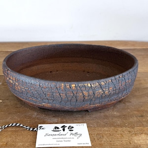 May include: A round, shallow ceramic bonsai pot with a dark, textured, crackled exterior finish revealing reddish-brown clay underneath. The interior is smooth and dark brown. A white rectangular tag with black text is attached by a string.
