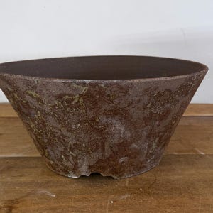 May include: A wide, conical ceramic bowl featuring a textured, mottled surface with deep brown and earthy bronze tones. The exterior displays a weathered, rustic finish with subtle variations in colour, while the interior is a dark, solid brown.