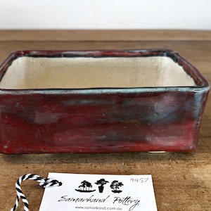 May include: A rectangular ceramic bonsai pot with a deep red, glossy exterior glaze and a smooth, off-white interior. A white tag attached with black and white twine displays the text Samarkand Pottery, www.samarkand.com.au, and 4457.