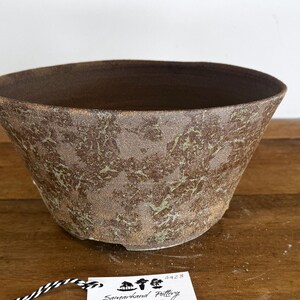 May include: A wide, conical ceramic bowl with a textured, mottled brown and greenish-grey exterior finish. The interior is a smooth, dark brown. A small white tag with the text Samarkand Pottery and the number 4428 is attached by a string.