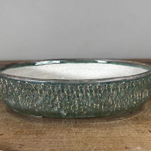 May include: A shallow, round ceramic bowl with a textured, moss-green exterior featuring vertical ridges and a smooth, speckled off-white interior. The rim is dark and defined, contrasting with the earthy green tones of the outer wall.