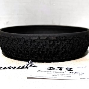 May include: A round, black ceramic bowl with a textured, patterned exterior. The bowl has a wide, shallow design. A tag with text is in the foreground. The bowl is likely handmade pottery.