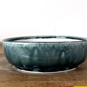 May include: A shallow, round ceramic bowl featuring a deep teal and dark blue reactive glaze. The exterior surface displays a glossy finish with subtle vertical drips and variations in color intensity, while the interior rim shows a lighter tone.