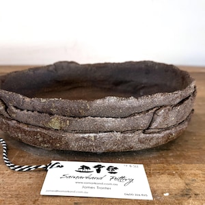 May include: A stack of three dark brown, textured ceramic bowls with irregular, rough edges. A white rectangular tag is attached to the bowls with a black and white striped string, featuring the text Samarkand Pottery, a website, and contact details.