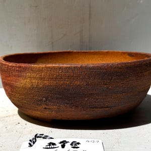 May include: An oval-shaped bonsai pot made from ceramic. The pot has a textured, earthy brown exterior and a lighter orange-brown interior. The design is simple and functional, suitable for bonsai cultivation.