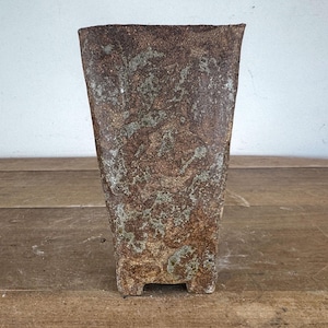 May include: A square-shaped ceramic vase with a rough, textured surface. The exterior features a mottled color palette of deep brown, rust, and muted green tones. The vessel has a wide, open top and a small, notched base that elevates it slightly.