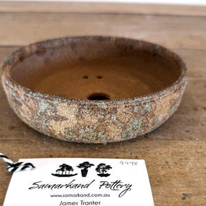 May include: A round, shallow ceramic bonsai pot with a textured, mottled brown and green exterior finish. The interior is a smooth, solid brown with a central drainage hole and two smaller holes. A white tag with black text reads Samarkand Pottery.