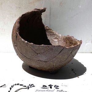 May include: A handcrafted, brown ceramic bowl with a unique, textured surface and a distinctive, curved rim. The bowl features a rough, organic design with visible cracks and a natural, earthy aesthetic. The base has three small feet.