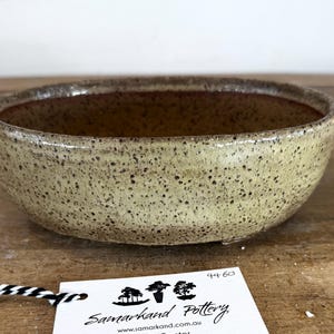 May include: A round ceramic bowl with a speckled light beige and tan exterior glaze and a smooth dark brown interior. A white rectangular tag is attached with black and white twine, featuring the text Samarkand Pottery and the number 44-60.
