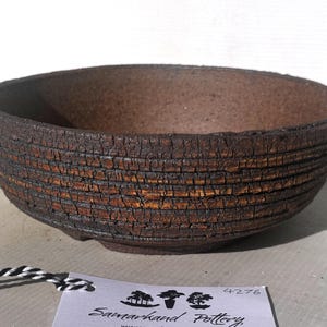 May include: A handcrafted, brown pottery bowl with a textured, layered design. The bowl has a wide, open shape and a dark, earthy color. The exterior features a rough, cracked texture, adding to its rustic appeal. The bowl is likely used for serving or display.