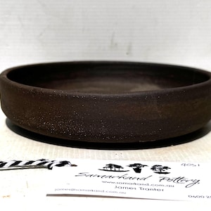 May include: A dark brown, round pottery bowl with a low profile. The bowl has a textured surface and a wide rim. A tag with the text "Samarkand Pottery" is visible. The bowl is likely handmade and suitable for serving or display.