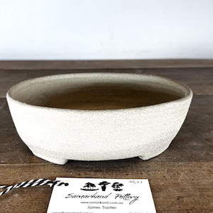 May include: A shallow, round ceramic bonsai pot with a textured, off-white matte finish. The bowl features a wide opening, a smooth interior, and small feet at the base. A white rectangular tag with black text reading Samarkand Pottery is attached.