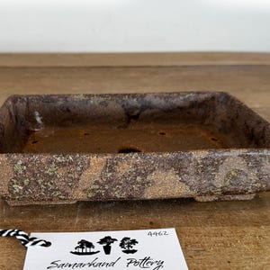 May include: A rectangular, brown, textured ceramic bonsai pot with a weathered, earthy finish. The pot features drainage holes at the bottom and sits on small feet. A white tag with the text Samarkand Pottery and the number 4462 is attached.