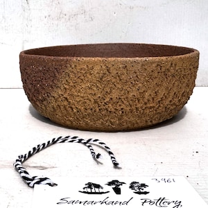 May include: A textured ceramic bowl with a rough, earthy exterior. The bowl has a wide, shallow shape with a dark brown interior and a lighter, tan-coloured exterior. A tag with the text "Samarkhand Pottery" is visible.