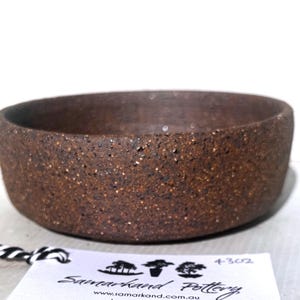 May include: A small, round, brown ceramic bowl with a textured surface. The bowl has a wide opening and a simple design. The bowl is likely handmade and suitable for serving food or as a decorative piece.