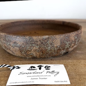 May include: A round, shallow ceramic bowl with a textured, mottled brown and earthy green finish. Attached is a white tag with black text reading Samarkand Pottery, www.samarkand.com.au, James Tranter, 4501, james@samarkand.com.au, and 0400 214 615.