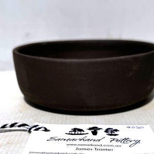 May include: A dark brown, circular ceramic bowl with a matte finish and straight sides. Attached to the bowl is a white rectangular tag featuring the text Samarkand Pottery, the website samarkand.com.au, and the name James Tranter.