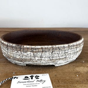 May include: An oval ceramic bonsai pot with a dark brown interior and a textured exterior featuring a white crackle glaze over a dark base. A white tag is attached, displaying the text Samarkand Pottery, a website, and the name James Tranter.