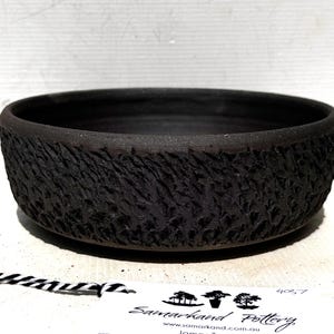 May include: A round, shallow black ceramic bowl with a heavily textured, rugged exterior surface. The interior of the bowl is smooth and matte black. A white paper tag with the text Samarkand Pottery is attached by a black and white string.