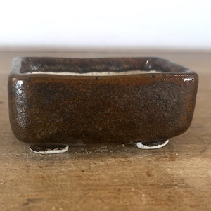May include: A rectangular ceramic bonsai pot with a dark brown, textured, glossy glaze on the exterior and a contrasting white interior. The vessel features four small, short feet at the base, providing elevation for the container on a flat surface.