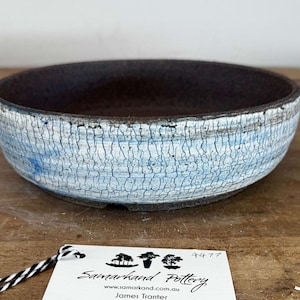 May include: A round ceramic bowl with a dark brown interior and a textured exterior featuring a white and light blue crackle glaze finish. A white rectangular tag is attached, displaying the text Samarkand Pottery, a website, and the number 4477.