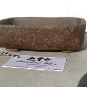 May include: A rectangular, brown stoneware planter with a textured surface. The planter sits on four small feet. A tag with text is attached to the planter.