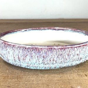 May include: A shallow, round ceramic bowl with a smooth, off-white interior. The exterior features a textured, drippy glaze in shades of light blue, lavender, and deep burgundy, creating a rugged, vertical, and layered appearance.