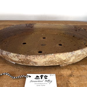May include: An oval ceramic bonsai pot with a textured, mottled brown and tan exterior finish. The interior base features five drainage holes. A white rectangular tag is attached with black and white string, displaying the text Samarkand Pottery.