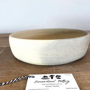May include: A shallow, round ceramic bowl with a textured, speckled off-white exterior and a smooth, light beige interior. A white rectangular tag is attached by a black and white string, featuring the text Samarkand Pottery, James Tranter.