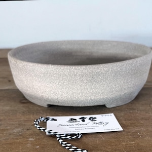 May include: A shallow, round, light gray ceramic bonsai pot with a textured, speckled finish and a wide opening. A white rectangular tag with black text reading Samarkand Pottery is attached by a black and white twisted string.
